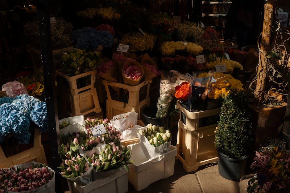 A vibrant outdoor flower market displaying an assortment of fresh flowers arranged in white plastic crates and black buckets. Prominent among these are pink and white tulips, blue hydrangeas, and yell