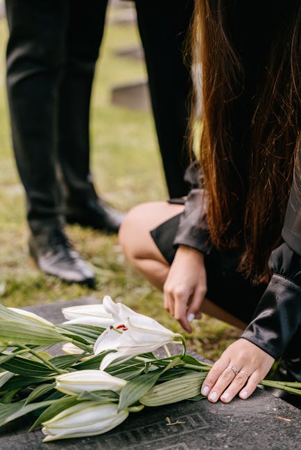 A person kneeling beside a floral arrangement of white lilies with long green leaves on a flat surface, possibly a gravestone, during a memorial or funeral setting. The individual’s hand gently rest
