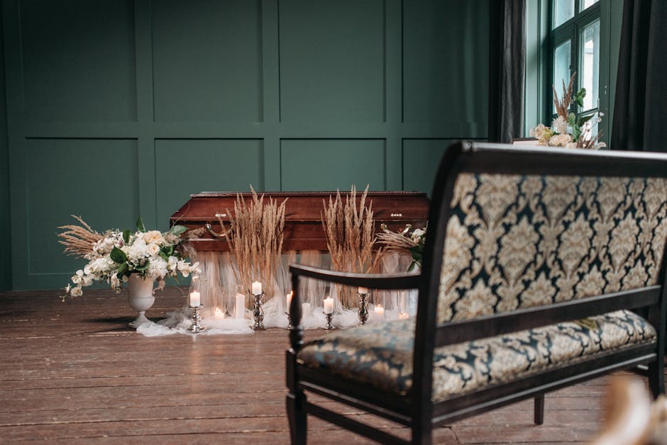 A floral arrangement placed on a wooden surface in front of a green paneled wall, featuring white and cream flowers with green foliage arranged in a white vase, complemented by tall, dried pampas gras
