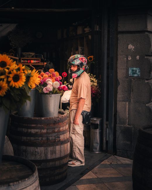 A person wearing a colorful helmet with a floral design stands beside large buckets filled with vibrant fresh flowers, including pink, purple, and peach blossoms, arranged in a rustic outdoor setting