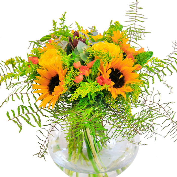 The essence of sun-drenched joy is poured into this showstopping bouquet-a living composition shaped by the spirit of Spitalfields floral artistry. Large, vibrant sunflowers take centre stage, their golden petals and dark velvet centres unfurling beside plush yellow roses in elegantly furled layers. Glossy red Hypericum berries catch the light and glimmer between tufts of feathery green fern and delicate filler, infusing energy and movement reminiscent of the bustling atmosphere outside Spitalfields Market Hall. Wisps of eucalyptus add hints of calming blue-green, pairing rustic charm with contemporary flair. Each flower's stem sits visible in a sparkling glass vase-every line and swirl on display, surrounded by fresh, crystalline water that keeps the arrangement vivid and alive. Light pours in and dances across the surfaces, enriching each hue and revealing the fine interplay between suede-like sunflowers and silken rose petals. To the nose, there's a delicate, cheerful fragrance suggestive of countryside fields brought into an East London home. Whether for a joyful birthday, a heartfelt anniversary, or as a bold centrepiece for local celebrations, this arrangement is a testament to Spitalfields' thriving creativity and the hands-on care of its neighbourhood florists.