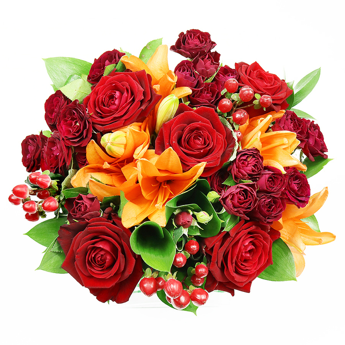 Captured from above on a stark white background, this richly hued floral arrangement forms a dense, circular statement of colour and craft. Several large deep red roses sit prominently at the heart of the design, their velvety, unfurling petals creating classic romantic focal points, while smaller, tightly clustered burgundy spray roses add concentrated pools of darker tone that deepen the overall jewel-like effect. Bright orange Asiatic lilies are woven through the composition, their trumpet shapes and visible stamens adding architectural lines and energetic bursts; a few remain in graceful bud, suggesting continued bloom and life. Bright red hypericum berries appear in small groups, their glossy spheres catching the light and providing a lively, almost festive texture against the softer petals. Lush green leaves - broad, shiny ruscus or salal - emerge between flowers to give the bouquet breath and to intensify contrast, creating a balanced and full profile. The soft, even lighting reveals the tactile interplay of velvet, satin and gloss, and the arrangement conveys warmth, celebration and a sense of occasion. This is the sort of bouquet a local florist on Commercial Street might assemble for a couple marking an anniversary by the old market stalls, or for a home in a Georgian mews near Christ Church; it feels both formal and approachable. Freshly conditioned stems and careful placement give the composition longevity and a composed beauty that suits romantic gestures, birthday tributes, or autumnal home decor.