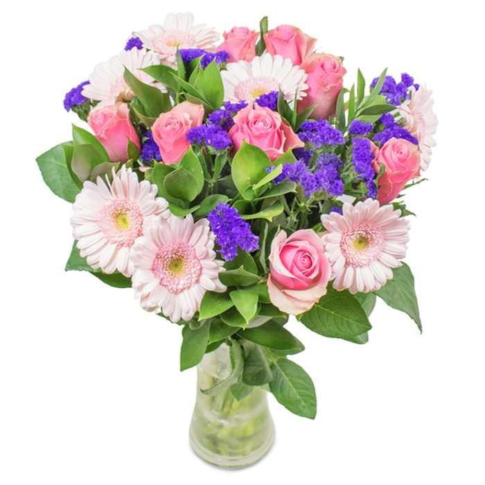 A striking floral arrangement presented in a transparent glass vase on a pristine white background, this bouquet captures the dynamic character of Spitalfields while staying true to classic beauty. At its core, seven to nine soft-to-medium pink roses-some petals still tightly closed, others just beginning to spiral open-draw in the gaze, their gentle pinks evocative of sunset over the market rooftops. Surrounding these, five to seven pale pink gerbera daisies bloom generously, each with a nearly white-painted centre that catches the light. Accentuating the composition, clusters of vivid purple statice bring a burst of deeper colour and textural intrigue-mirroring the playful energy found along Commercial Street's vibrant storefronts. The base of rich, emerald-green foliage lends a lush frame and airy abundance, carrying a subtly herbal scent reminiscent of newly cut stems at the morning flower stalls. Every stem is arranged in crystal-clear water within the vase, its radiance suggesting freshness and attentive care by local Spitalfields florists. Whether chosen as a cheerful birthday surprise, a thoughtful anniversary gesture, or a welcoming addition to a bustling studio, this bouquet overflows with feelings of joy, celebration, and deep affection-perfect for everywhere from Artillery Lane offices to Market Street homes.
